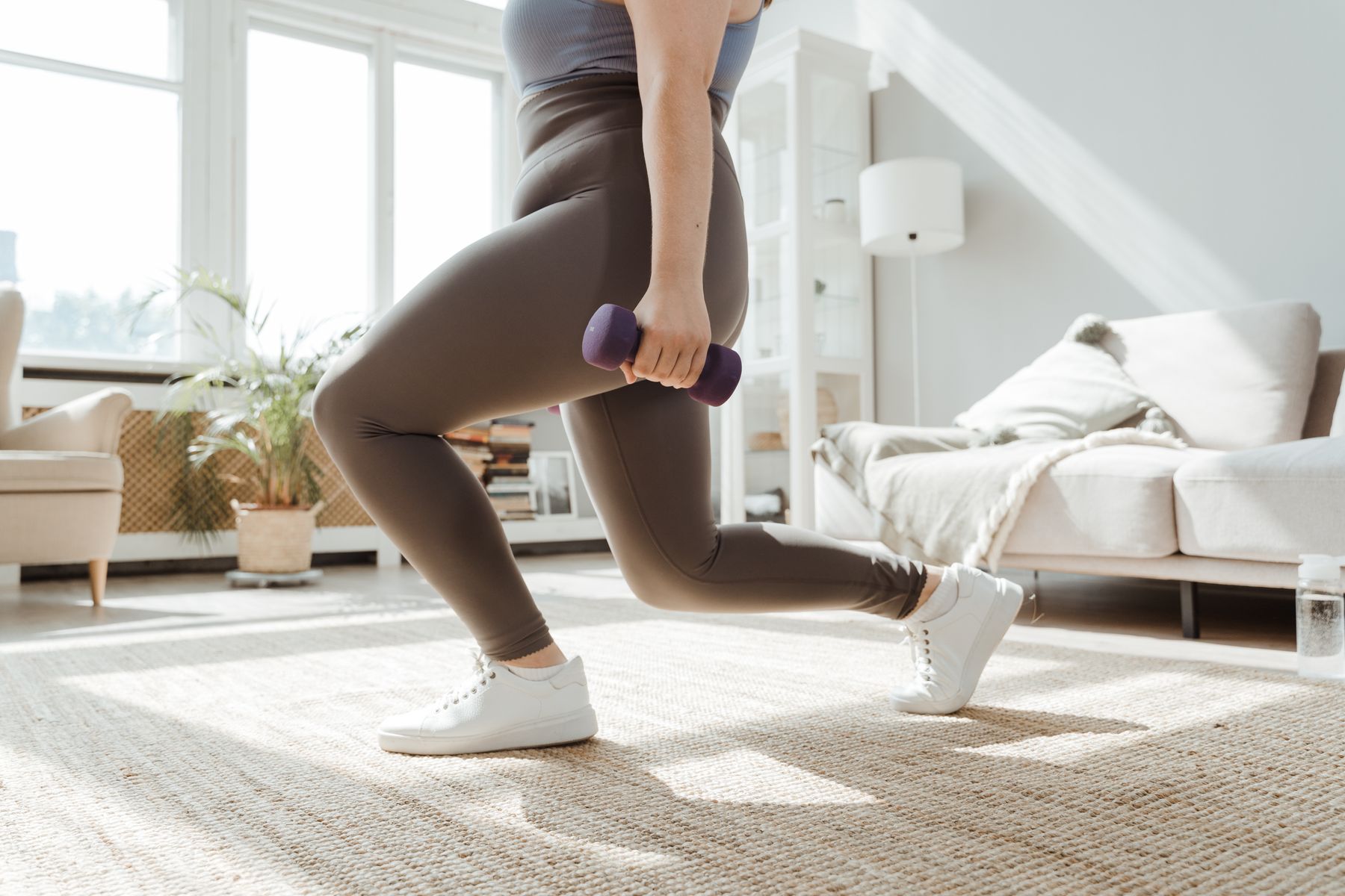 Close-up of an adult performing a dumbbell lunge in a bright home training space.