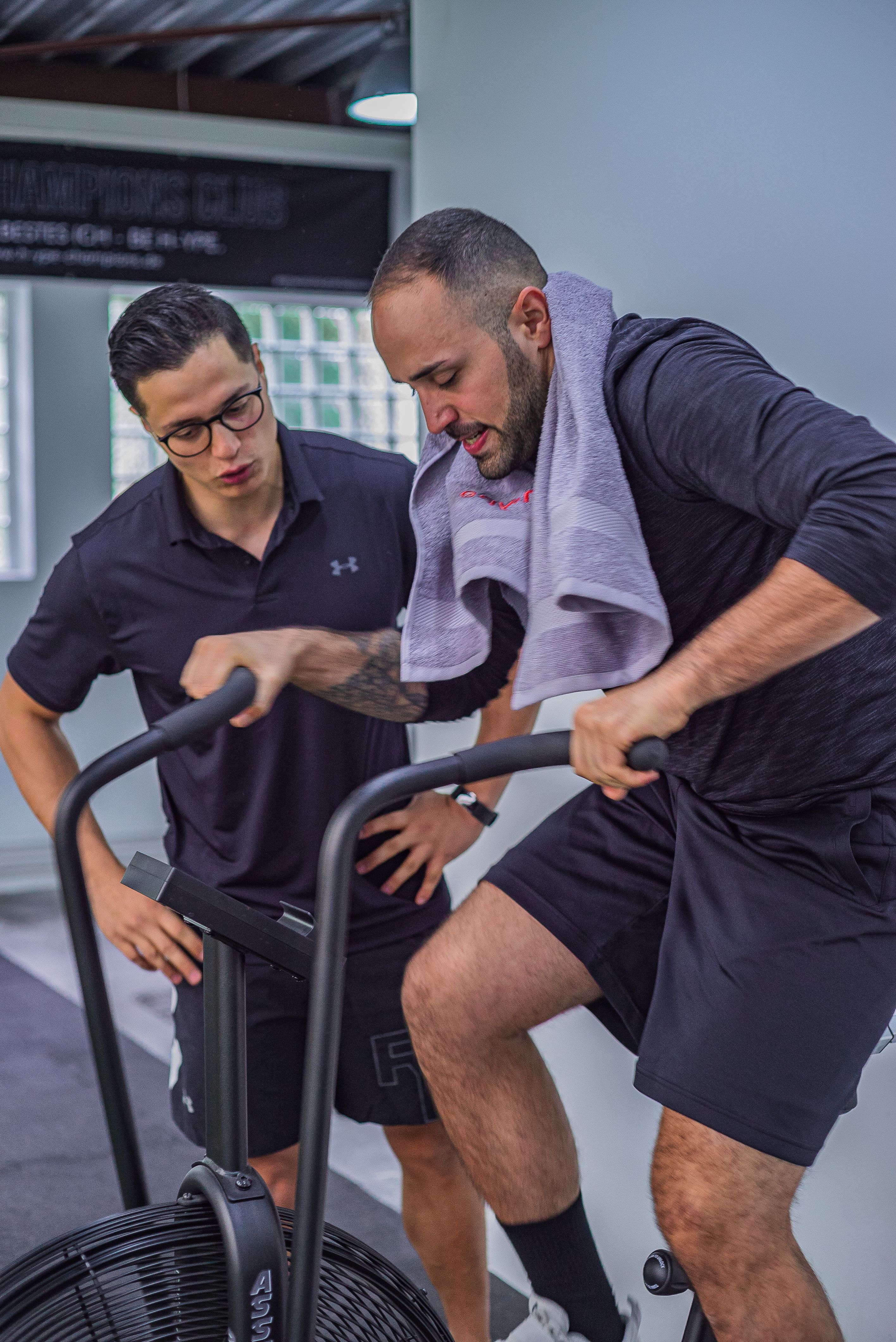 Adult riding an air bike while a trainer monitors effort in a gym.
